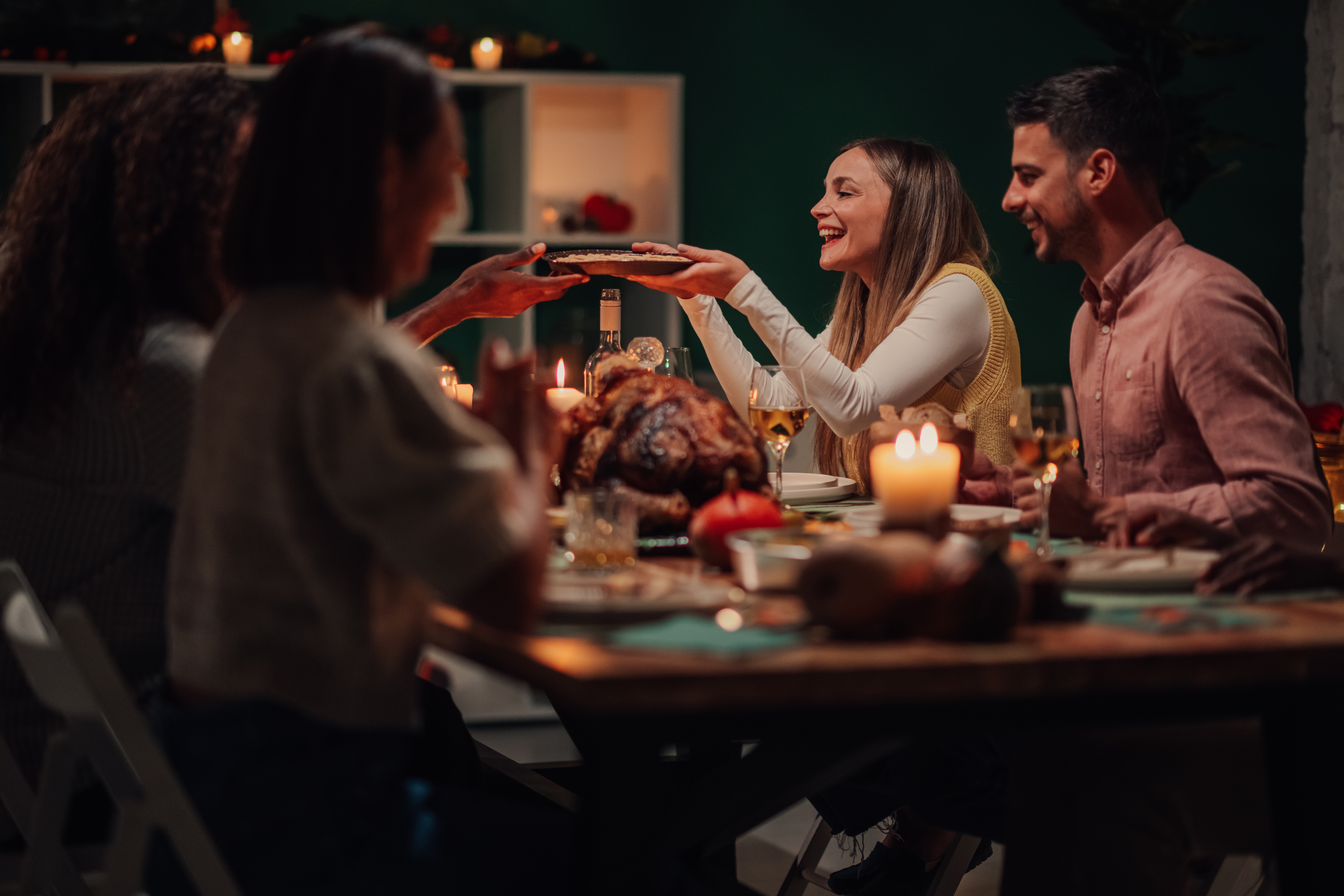 Group of friends sharing a festive meal at a candlelit dinner table during a cozy Thanksgiving celebration, smiling and passing food.