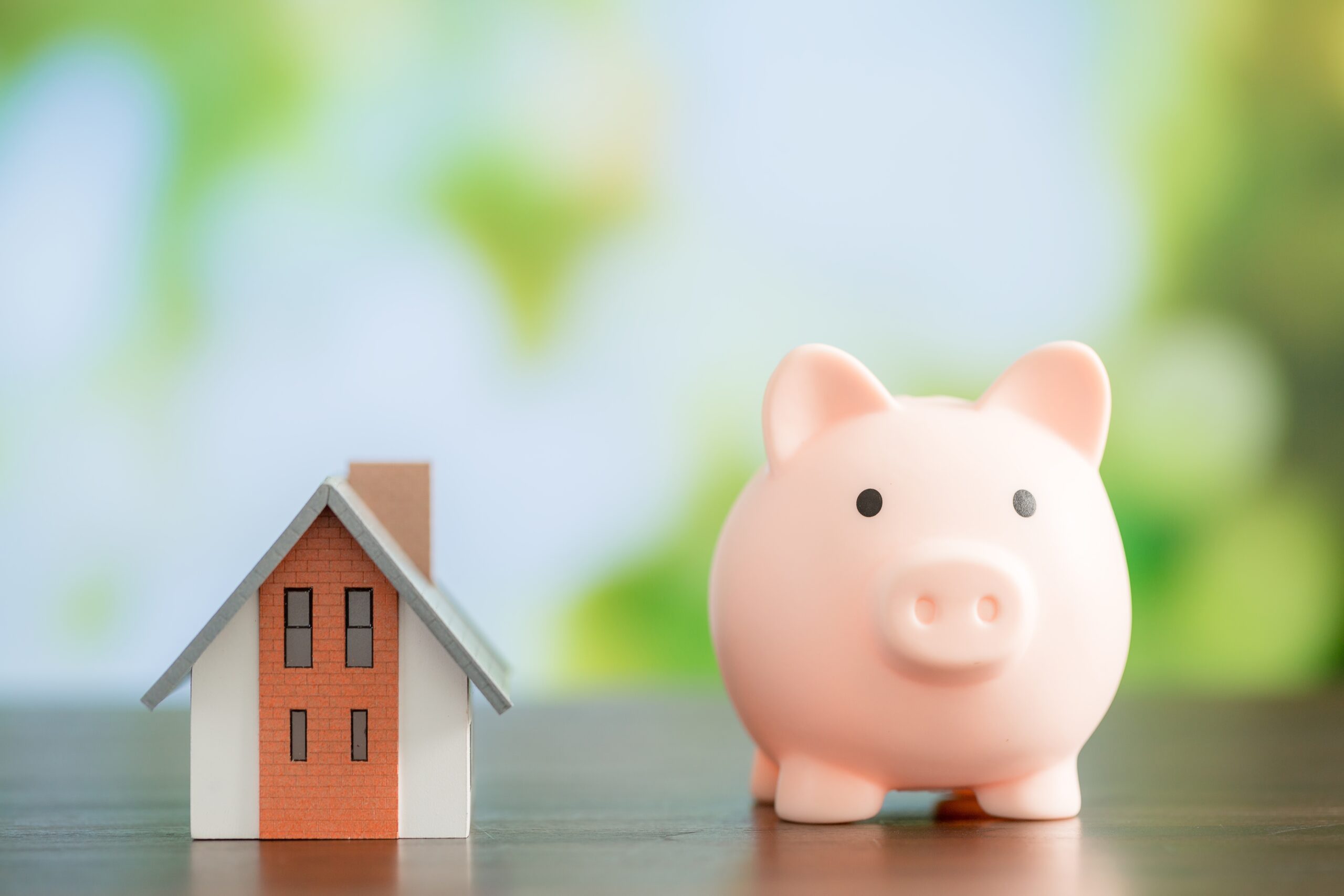 Pink piggy bank next to a small model house on a table, symbolizing household savings and personal finance.